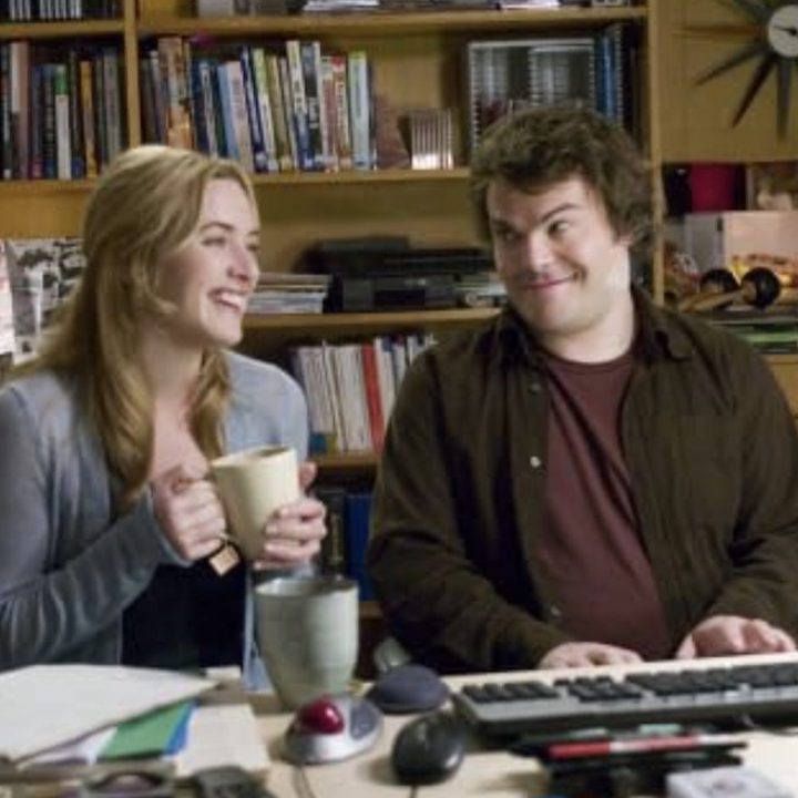 A woman holding a coffee cup sits down to a man at a keyboard.