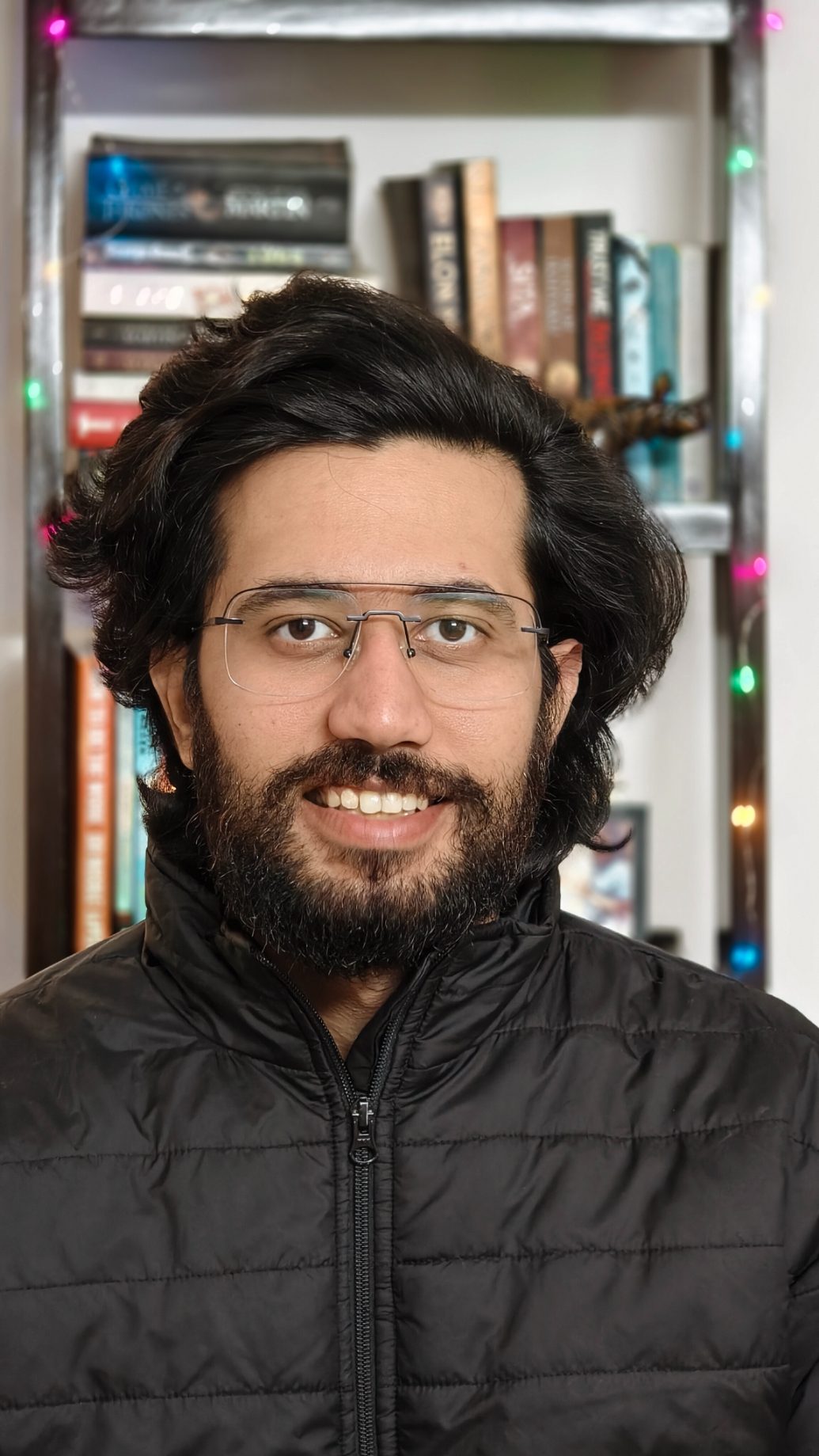 Portrait of a man in black bomber jacket in front of a book rack with fairy lights shot on the Vivo X100 Zeiss camera.
