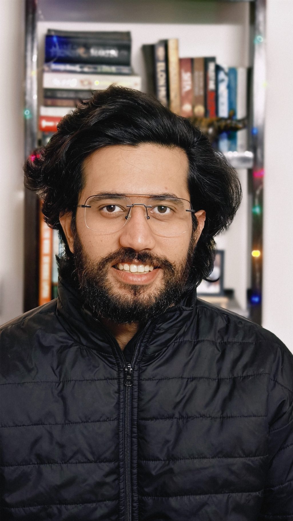 Portrait of a man in black bomber jacket in front of a book rack with fairy lights shot on the Vivo X100 Zeiss camera.