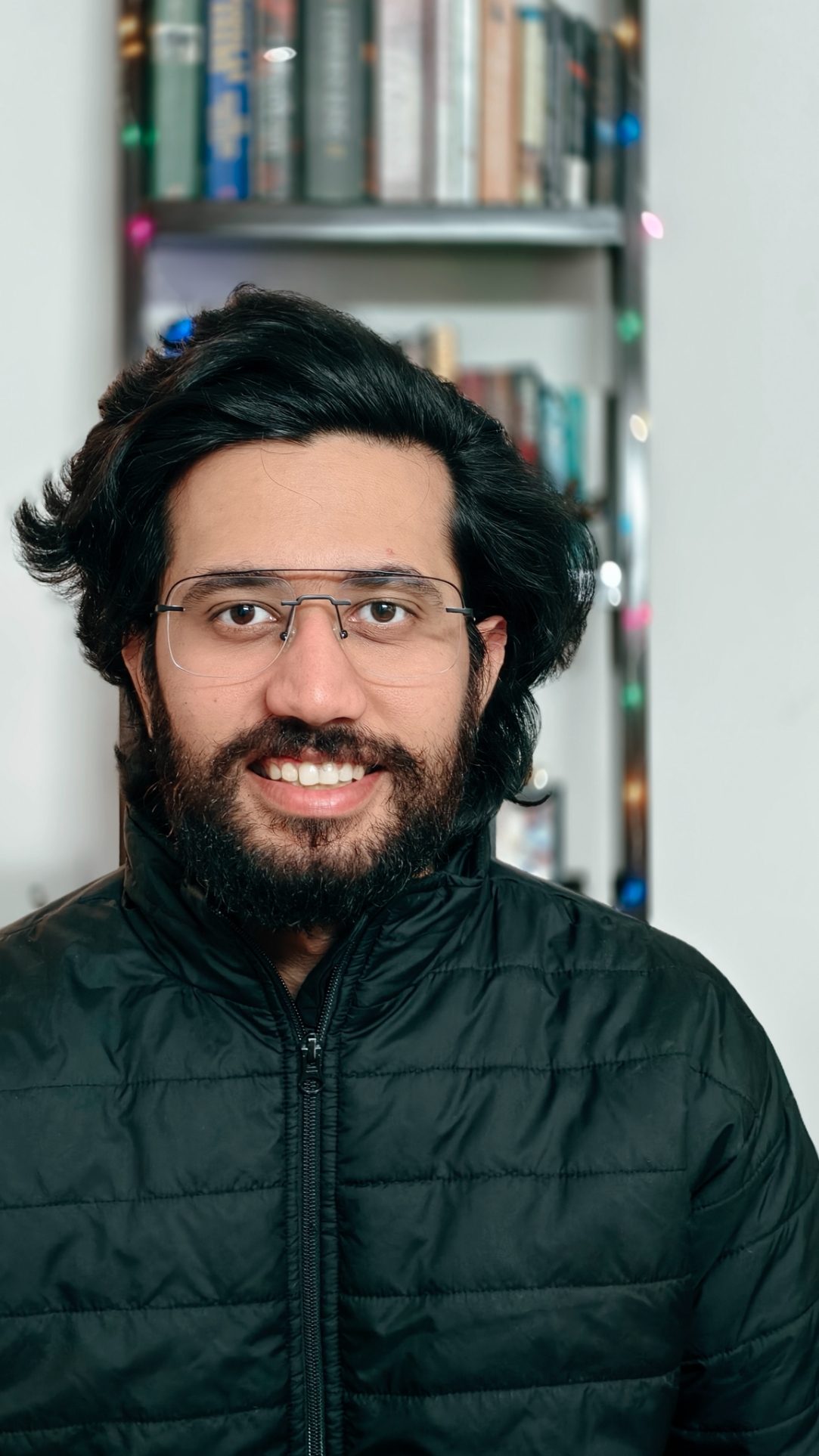 Portrait of a man in black bomber jacket in front of a book rack with fairy lights shot on the Vivo X100 Zeiss camera.
