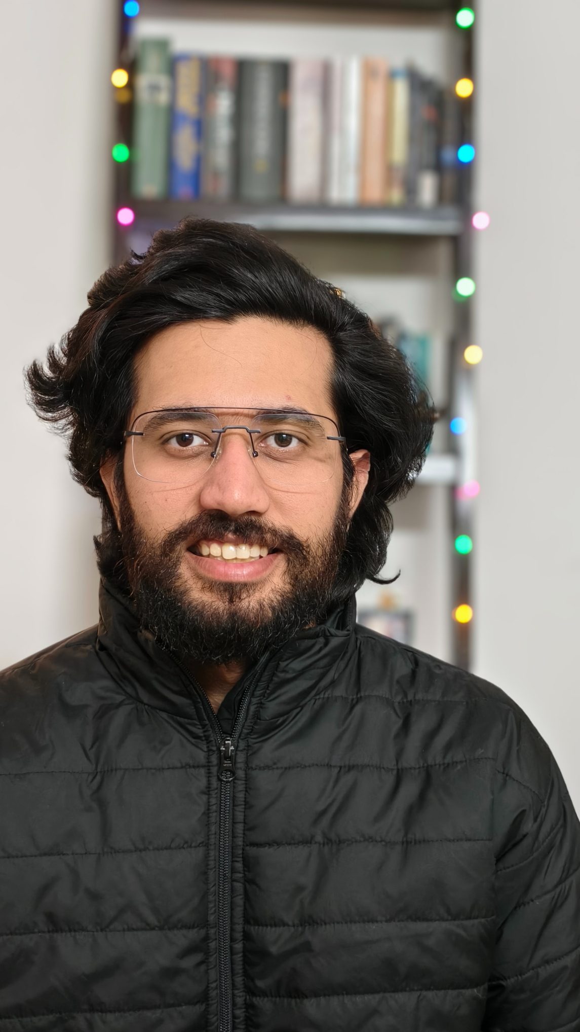 Portrait of a man in black bomber jacket in front of a book rack with fairy lights shot on the Vivo X100 Zeiss camera.