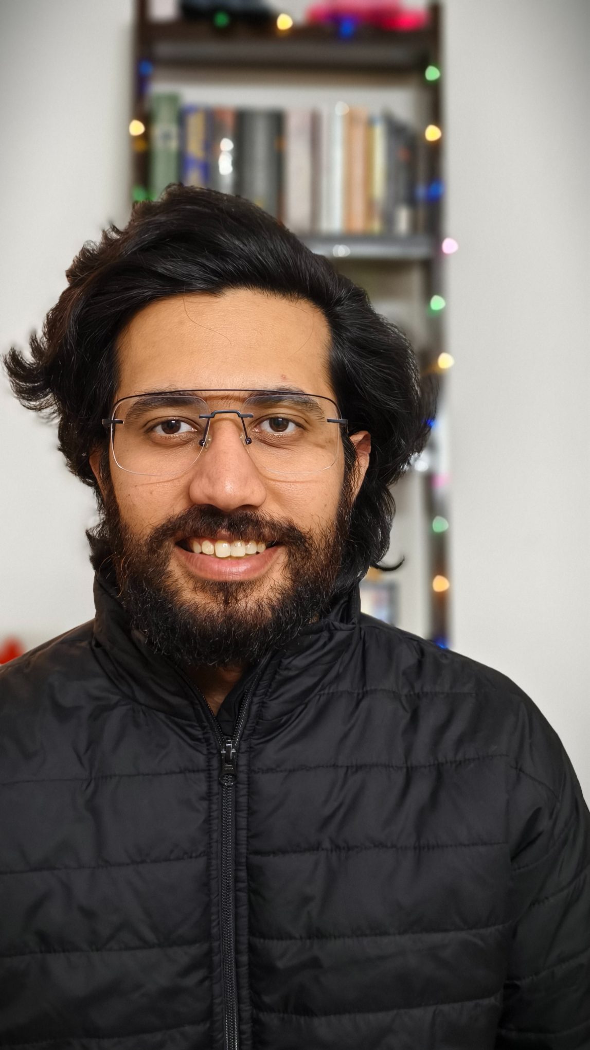 Portrait of a man in black bomber jacket in front of a book rack with fairy lights shot on the Vivo X100 Zeiss camera.