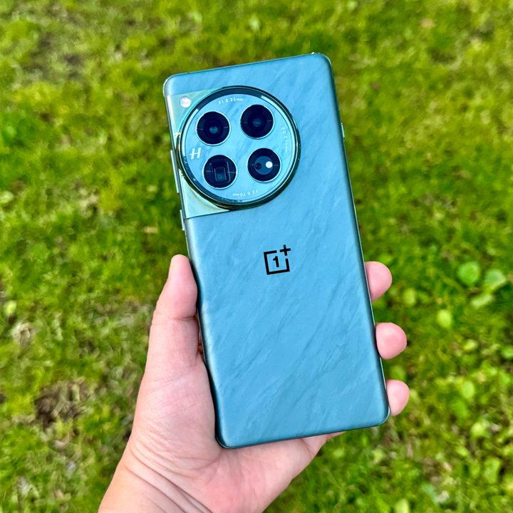 OnePlus 12 Flowy Emerald held in hand against grass background.