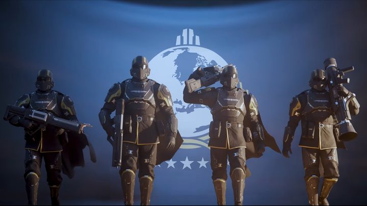 Four soldiers walk in front of a blue Super Earth flag in the Helldivers 2 trailer.