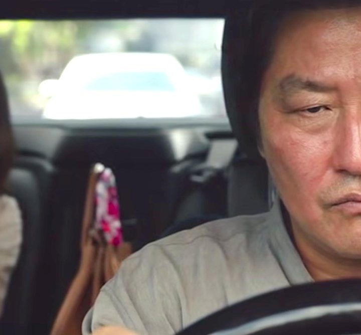 Song Kang-ho grimaces behind a steering wheel as Cho Yeo-jeong talks obliviously on the phone in the backseat in a still from the movie Parasite.