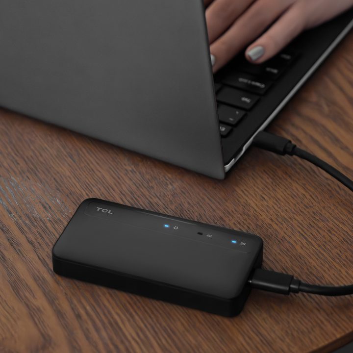 TCL Linkkey IK512 5G dongle on a table plugged into a laptop that someone is typing on.