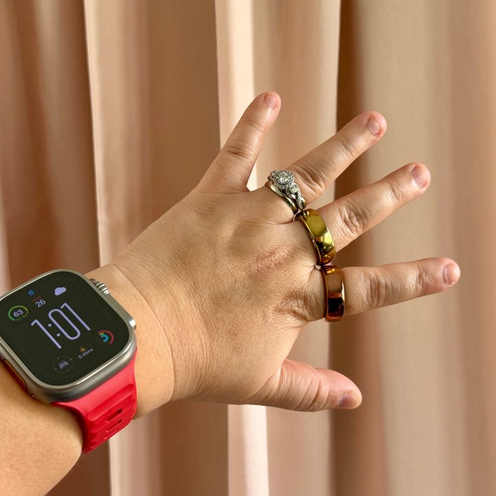 An Apple Watch Ultra with red strap on wrist with a Movano Evie and Oura Ring on fingers.