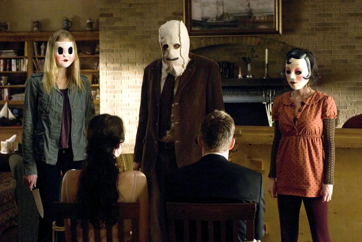 Three masked figures stand in front of a couple in The Strangers.