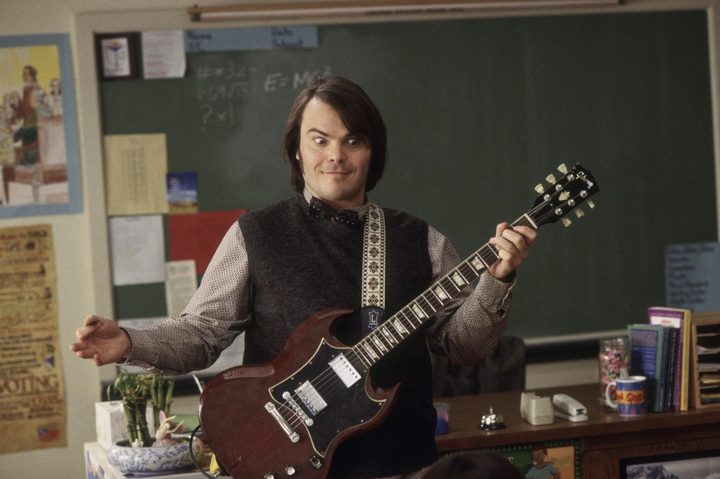 Jack Black holds a guitar in front of the room.