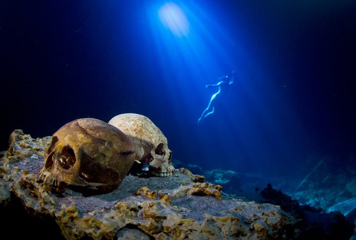 Mayan skulls in a sand with a swimmer behind it.