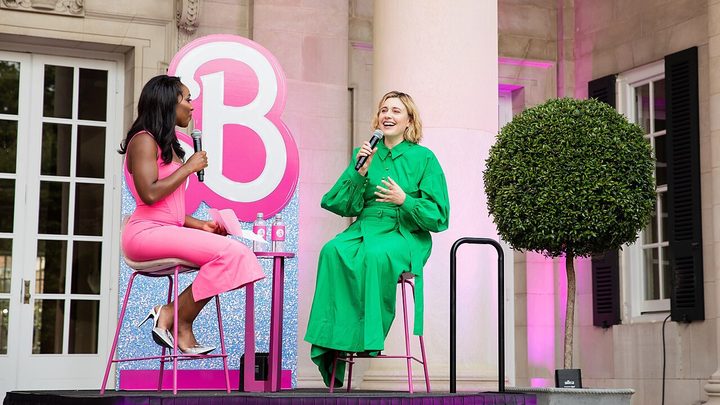 Greta Gerwig sitting with an interviewer, a Barbie backdrop in the background, laughing and speaking into a mic.