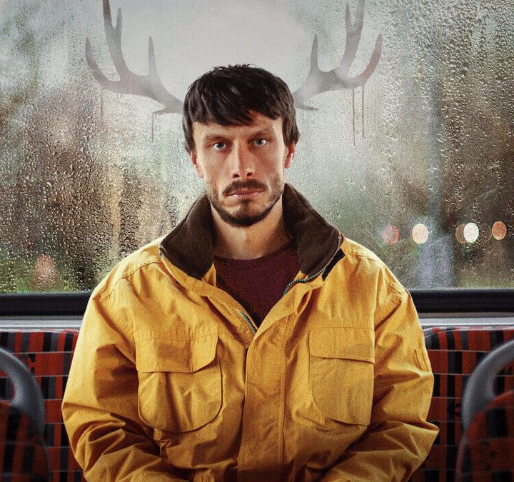 Richard Gadd in Baby Reindeer sitting on a bus with a yellow coat staring forward, the image of deer ears on either side of him.