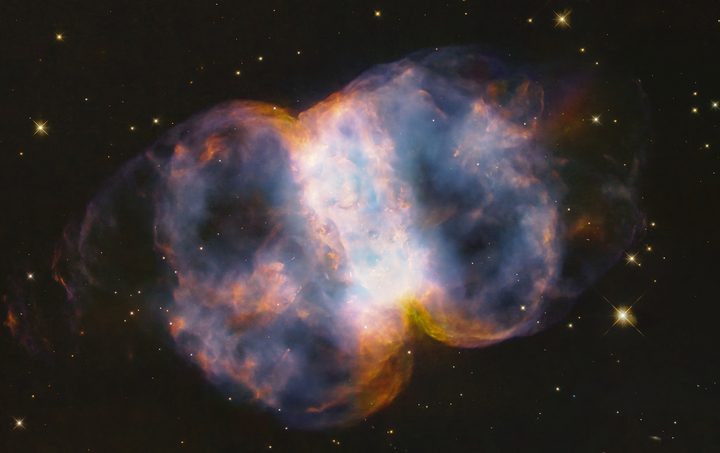 In celebration of the 34th anniversary of the launch of NASA’s legendary Hubble Space Telescope, astronomers took a snapshot of the Little Dumbbell Nebula, also known as Messier 76, or M76, located 3,400 light-years away in the northern circumpolar constellation Perseus. The name 'Little Dumbbell' comes from its shape that is a two-lobed structure of colorful, mottled, glowing gases resembling a balloon that’s been pinched around a middle waist. Like an inflating balloon, the lobes are expanding into space from a dying star seen as a white dot in the center. Blistering ultraviolet radiation from the super-hot star is causing the gases to glow. The red color is from nitrogen, and blue is from oxygen.