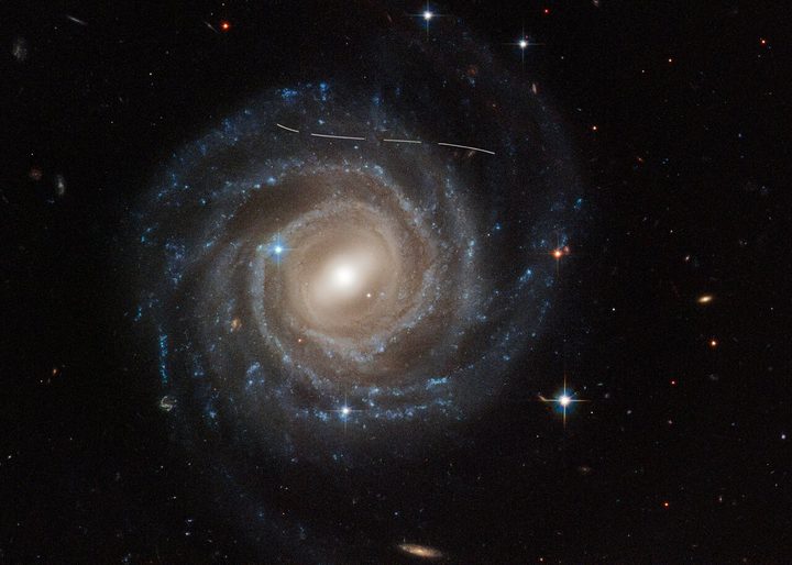 This NASA/ESA Hubble Space Telescope image of the barred spiral galaxy UGC 12158 looks like someone took a white marking pen to it. In reality it is a combination of time exposures of a foreground asteroid moving through Hubble’s field of view, photobombing the observation of the galaxy. Several exposures of the galaxy were taken, which is evidenced by the dashed pattern.