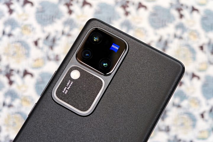 Close of a gray Vivo V30 Pro's camera module with T* Zeiss lens coating held against a printed cloth.