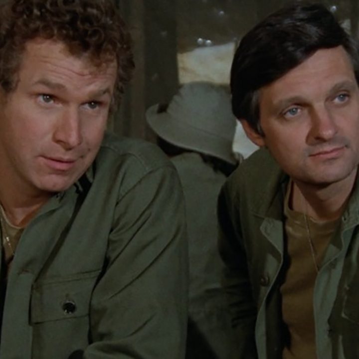 Two men sitting together, both wearing green army surgical uniforms in a scene from M*A*SH.