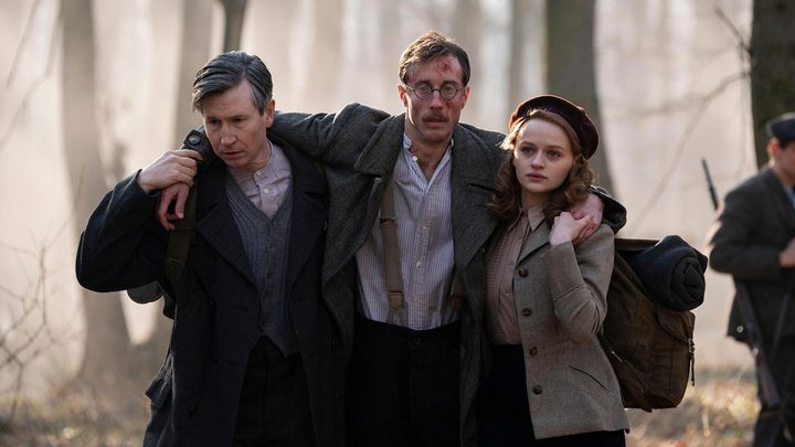 A young man and woman carrying an older, injured man, his arms around their shoulders on either side, in a scene from We Were the Lucky Ones.