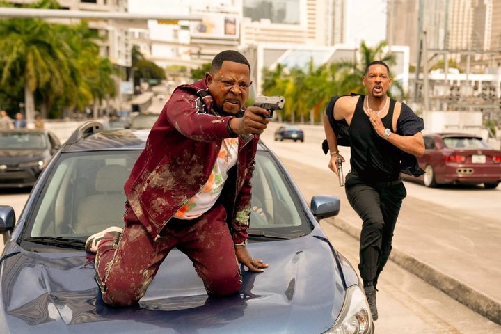 Martin Lawrence sits on a car hood while Will Smith runs next to him in Bad Boys: Ride or Die.