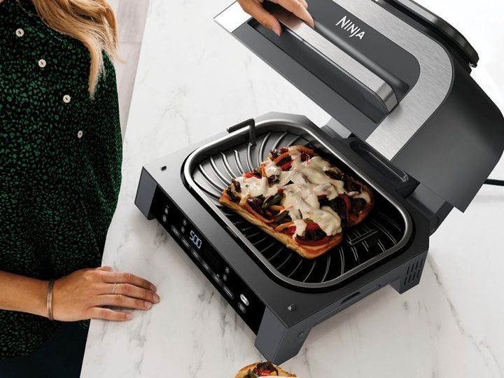 Lifting the lid of a Ninja Foodi XL 6-in-1 indoor grill to reveal a pizza cooking.