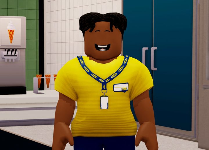 A smiling worker inside the Roblox Ikea experience. He's wearing a yellow shirt and has an Ikea lanyard with a key card around his neck.
