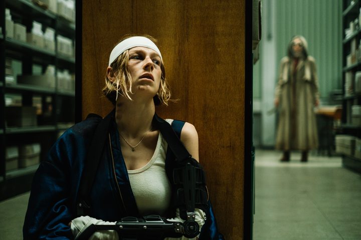 An injured woman sits up against a shelf as another woman stands down the hallway.