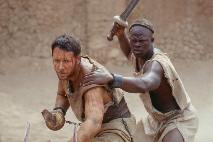 Russell Crowe points a sword as a man stands behind him with one.