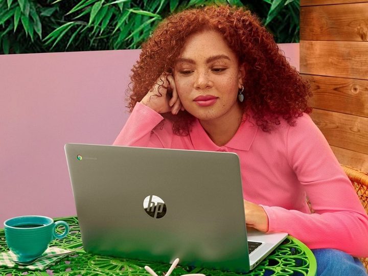 A woman using a HP Chromebook laptop while sitting at a bright green table with a cup of coffee, all in front of a colorful background.