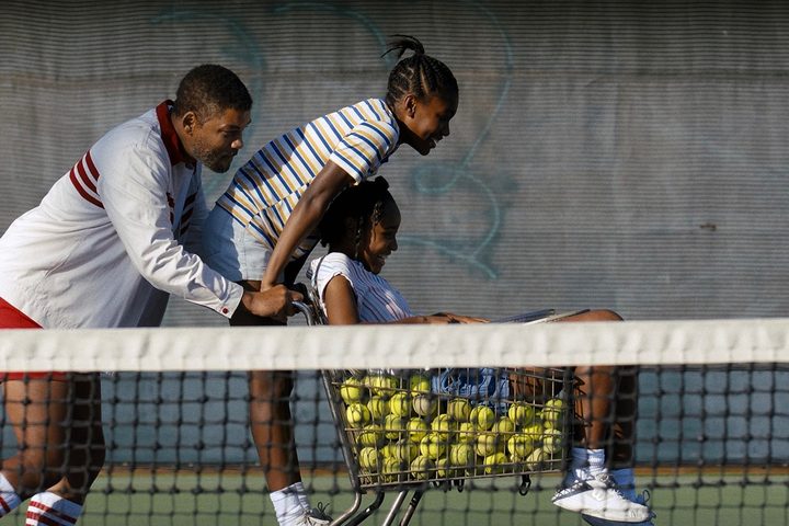 A man pushes a shopping cart of tennis balls with his two girls holding onto the cart.