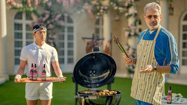 Jeff Goldblum barbecuing with an apron, another man holding a tray beside him in a scene from Kaos on Netflix.