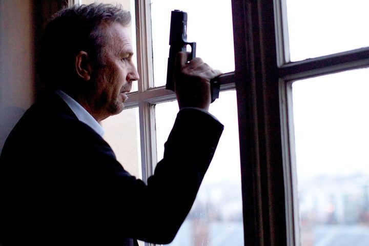 A man with a gun leans against a window in 3 Days to Kill.