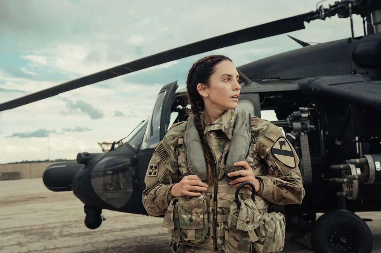 A soldier holds onto her pack as she stands in front of a helicopter.