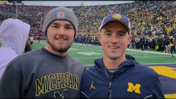 Two men pose for a picture in front of a football field.
