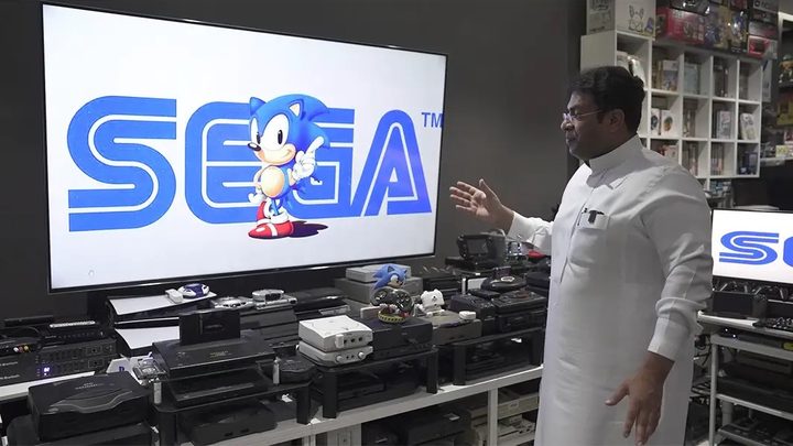 Ibrahim Al-Nasser standing in front of his console setup. There are a ton of consoles lined up in front of a TV. Sonic and the Sega logo are on the TV