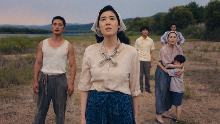 A group of people led by a woman with a kerchief around her hair look up to the sky in a scene from Pachinko.