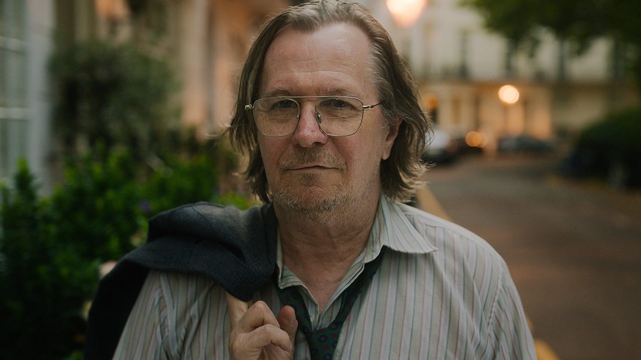 Gary Oldman smiling with his jacket draped over his shoulder in Slow Horses on Apple TV+.