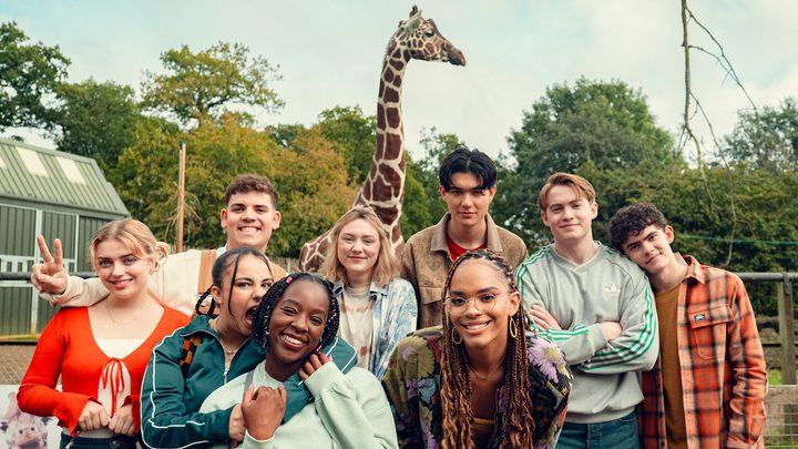 The cast of Heartstopper Season 3 smiling for a photo in front of a giraffe.