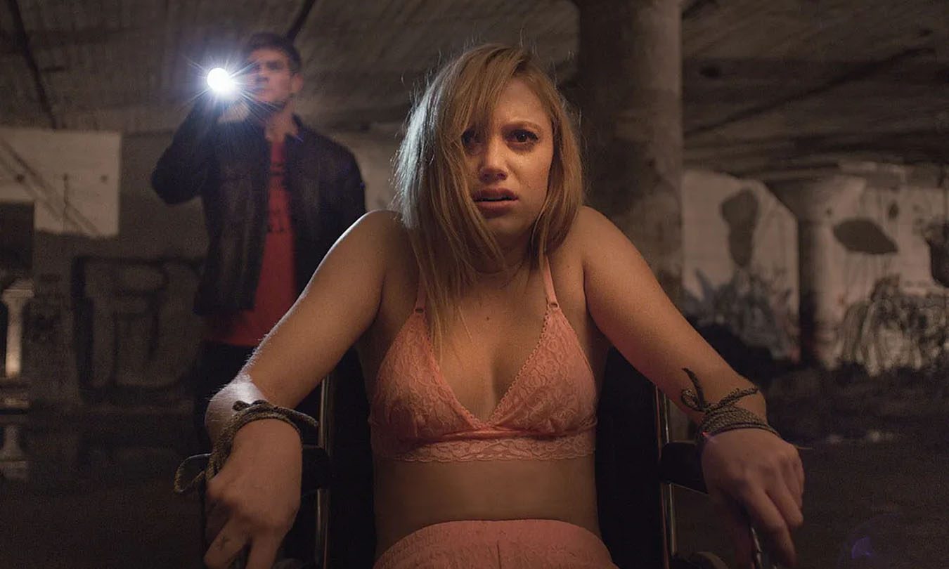 Maika Monroe bound to a chair with a man shining a flashlight behind her in It Follows.