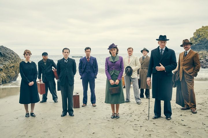 A group of people stand on a beach in And Then There Were None.