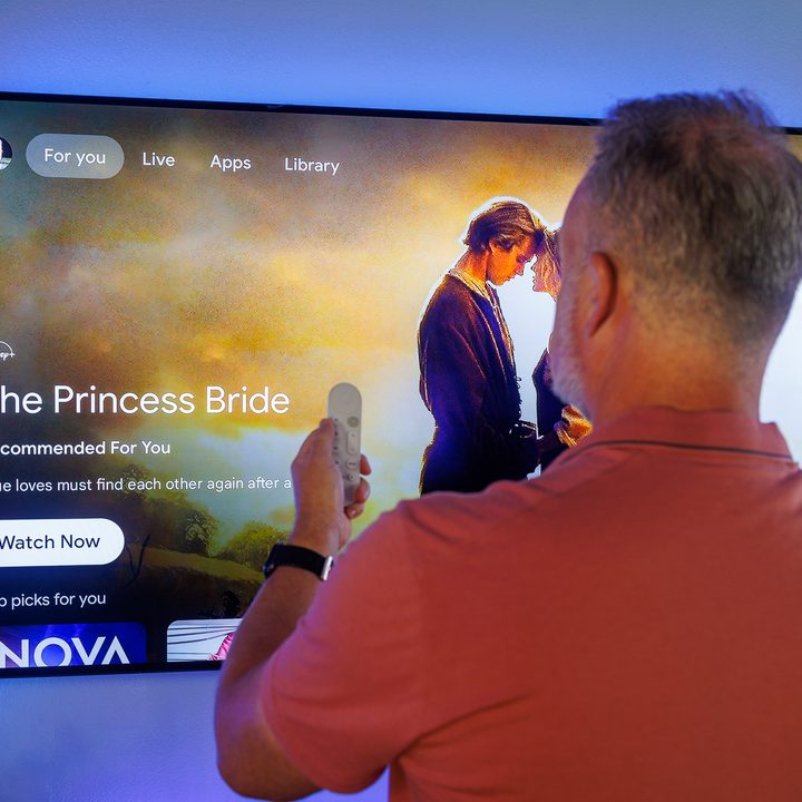 An aging journalist stands before the Google TV home screen on a television.