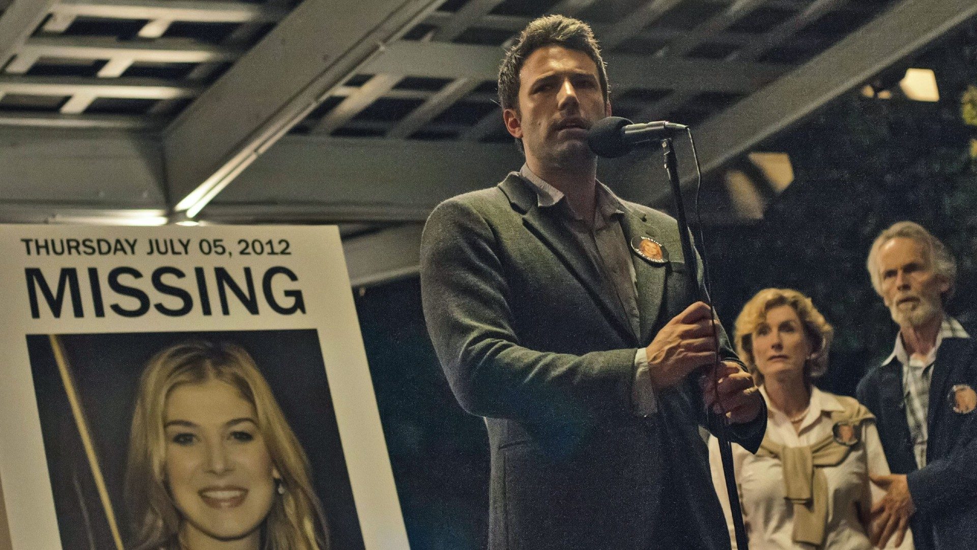 Ben Affleck stands in front of a blown-up photo of Rosamund Pike, holding a microphone, in a still from the movie Gone Girl.