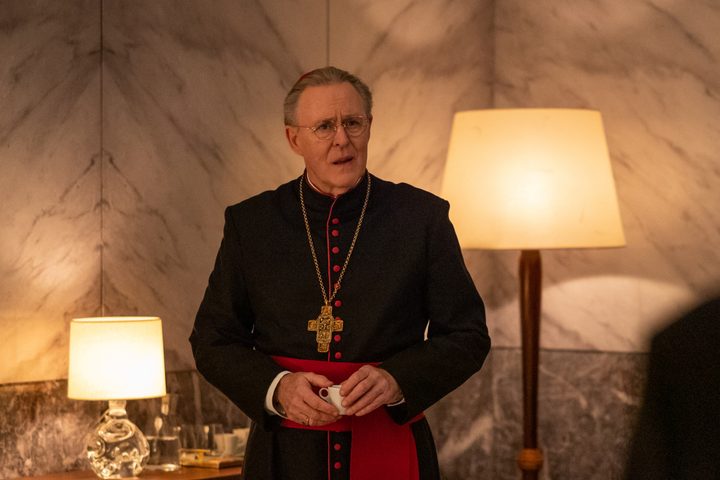 John Lithgow stands between two lamps in Conclave.