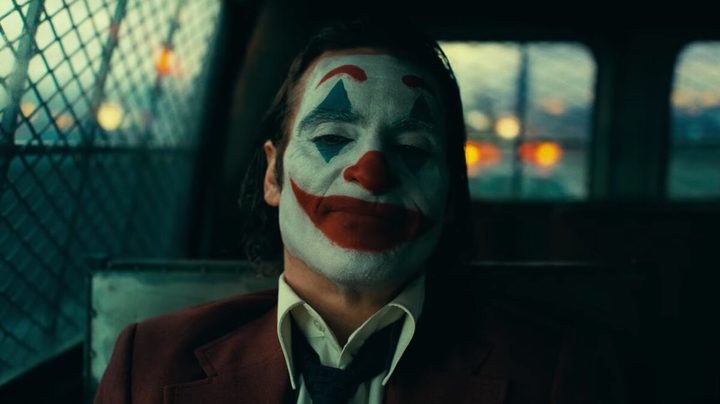 Joaquin Phoenix in full clown makeup sits in the back of a police cruiser in a still from the movie "Joker: Folie à Deux."