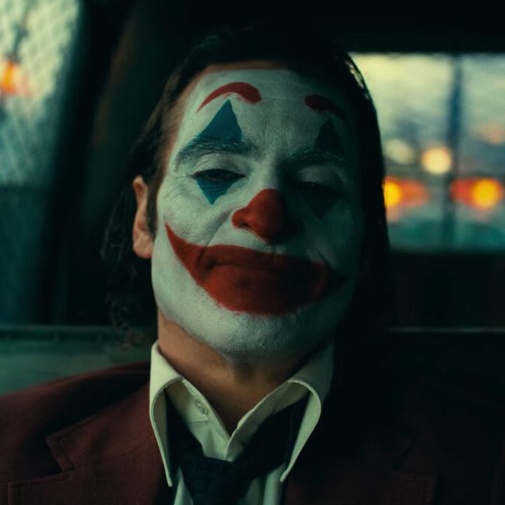 Joaquin Phoenix in full clown makeup sits in the back of a police cruiser in a still from the movie "Joker: Folie à Deux."