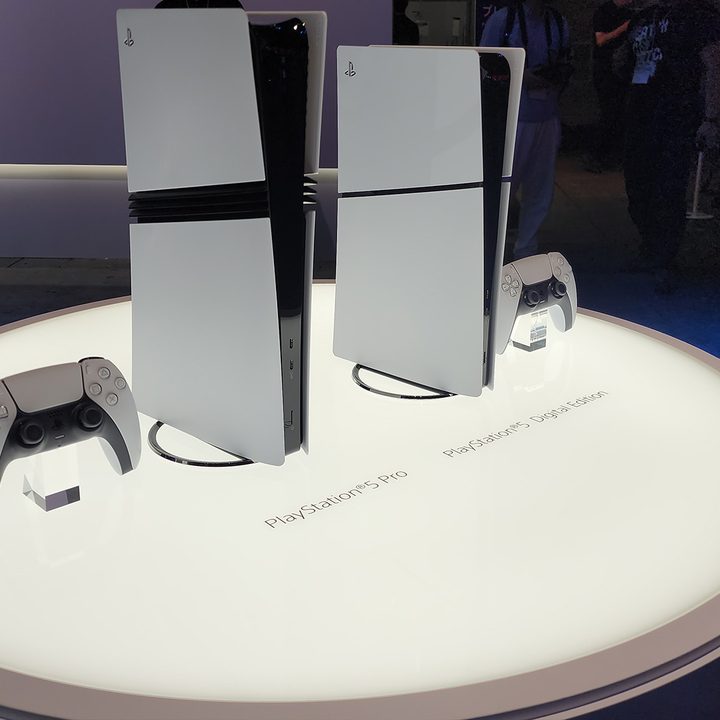 A lit table holds a PS5 Pro, and a PS5 all digital, the former standing noticeably taller.