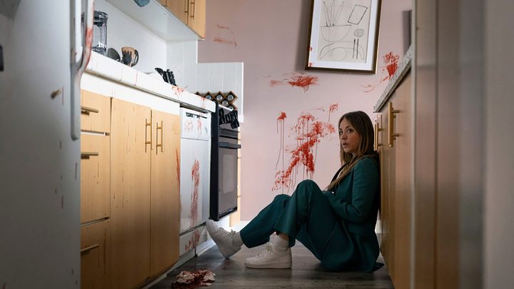 Ava sits on the floor in a kitchen, blood splattered on the way behind her, in Based on a True Story.