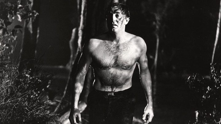 A black and white image of a man without a shirt in the middle of the forest looking worried.