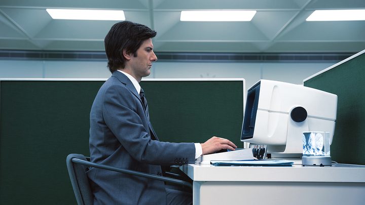 A side view of Mark at his desk in the office working on the computer in a suit in Severance on Apple TV+.