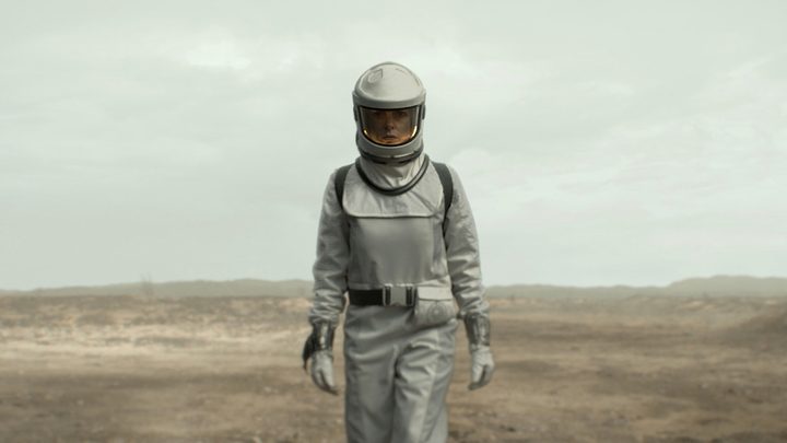 Juliette walking outside in an airtight uniform, a desolate landscape behind her in Silo.