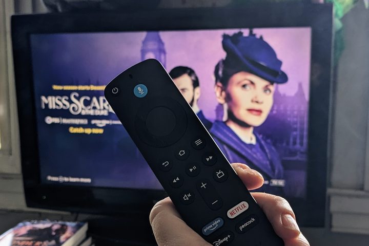 An Amazon Fire TV remote is held up in front of a TV. In the background a show is obscured by the remote.
