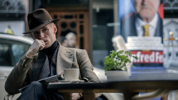 Eddie Redmayne in a hat sitting on a patio outside with his hand to his mouth, looking pensive in The Day of the Jackal.